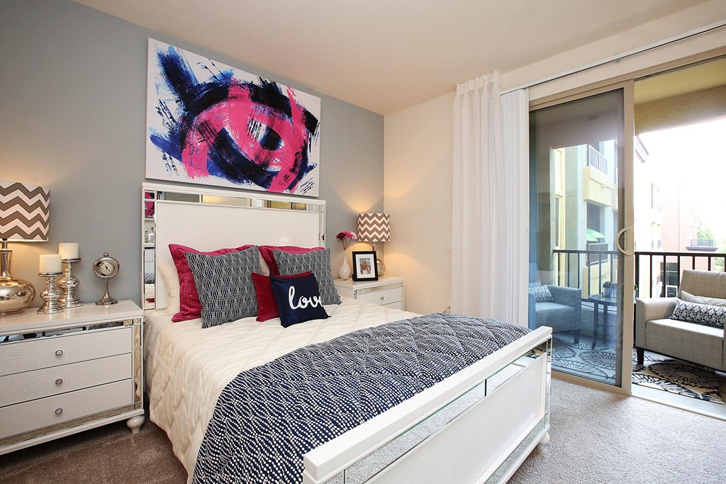 A bedroom with a large bed, a painting above it, and a chair in the doorway at The Kitt at Warner Center Apartments, Woodland Hills, CA