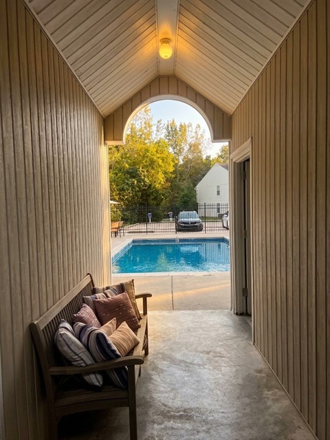 A wooden bench with cushions is placed in a hallway with a pool visible through an archway.