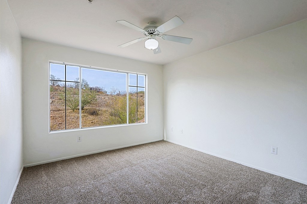 A room with a carpet floor, a ceiling fan, and a window showing a desert view.