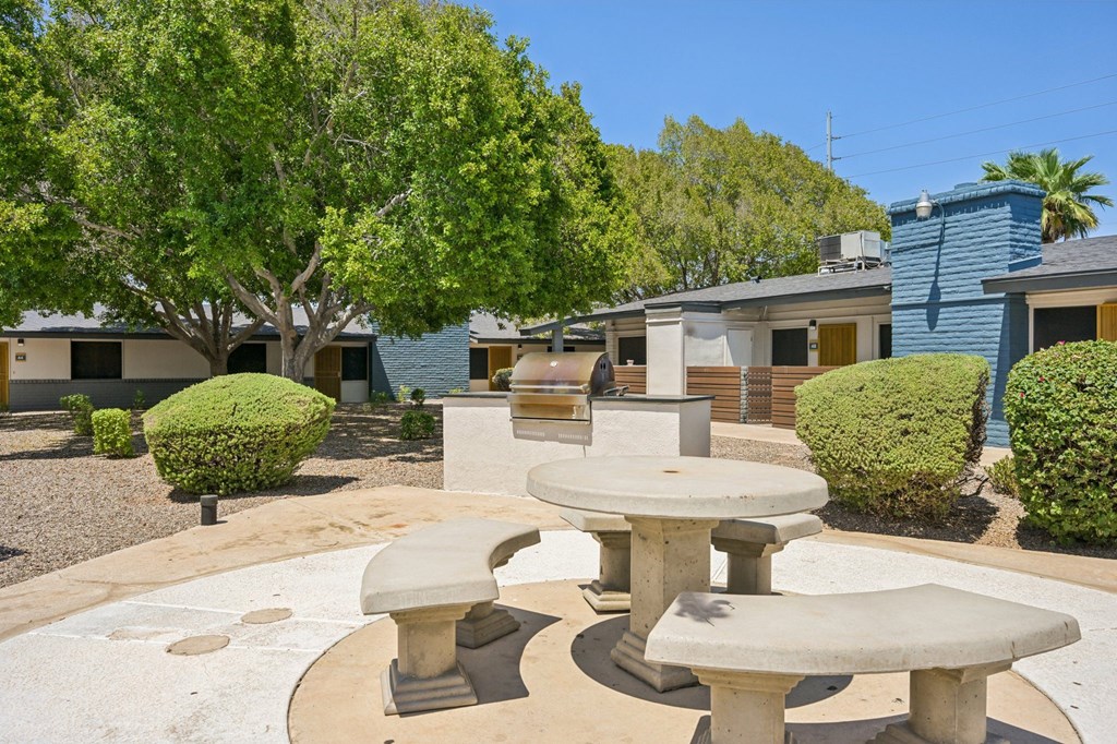 A concrete table and benches are in the foreground of a courtyard with houses and trees in the background.