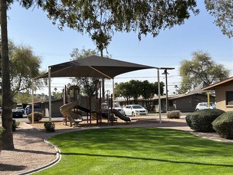 A playground with a slide and a canopy is surrounded by a grassy area and a sidewalk.