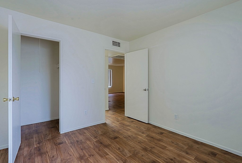 A room with white walls and wood flooring with a door on the left and a hallway on the right.