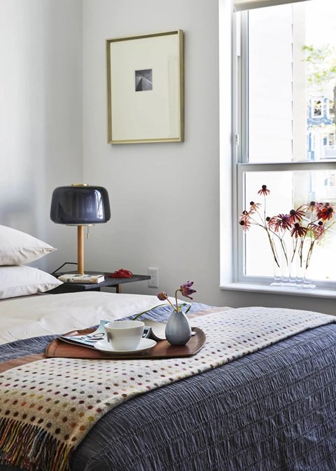 A bedroom with a bed, a lamp, a tray with a cup and a vase, and a window with a view of the outside.