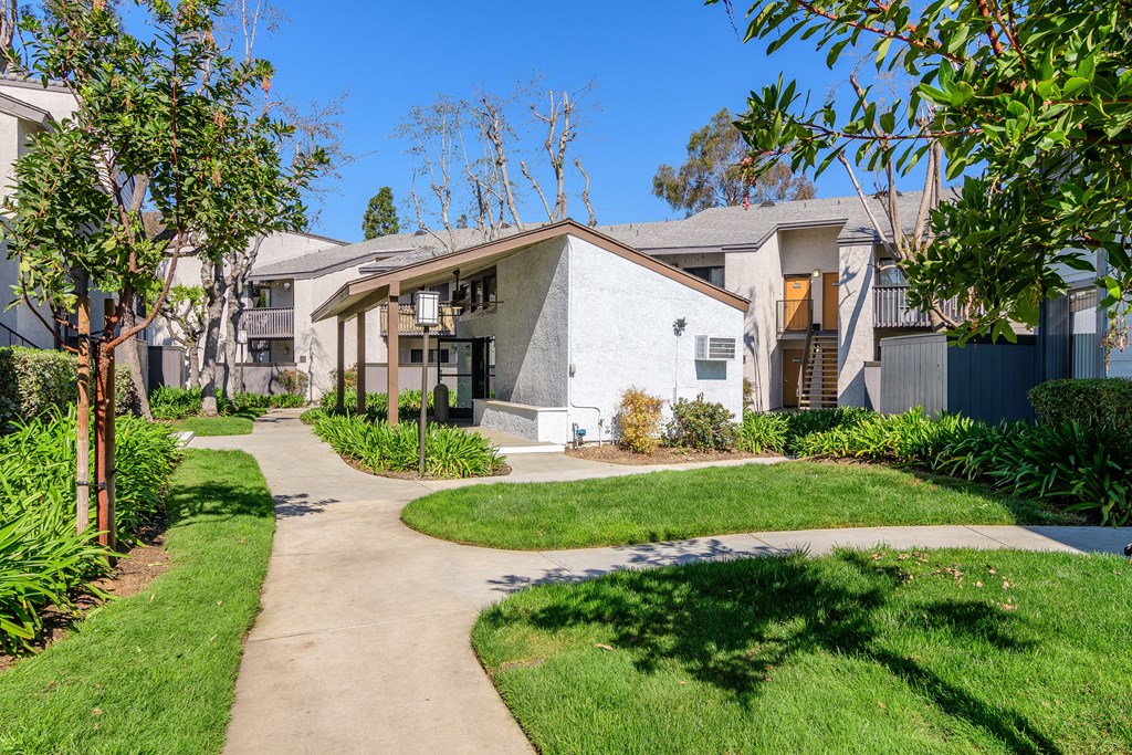 A white house with a grey roof is surrounded by greenery and a concrete pathway.