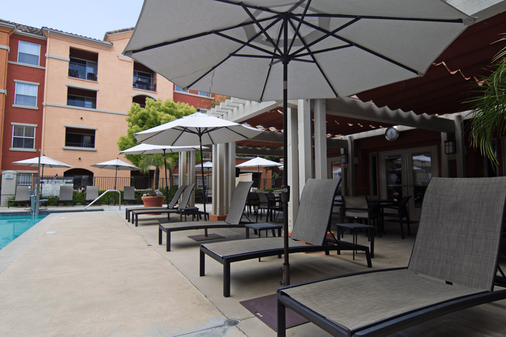A patio with chairs and umbrellas is surrounded by a pool and apartment buildings.