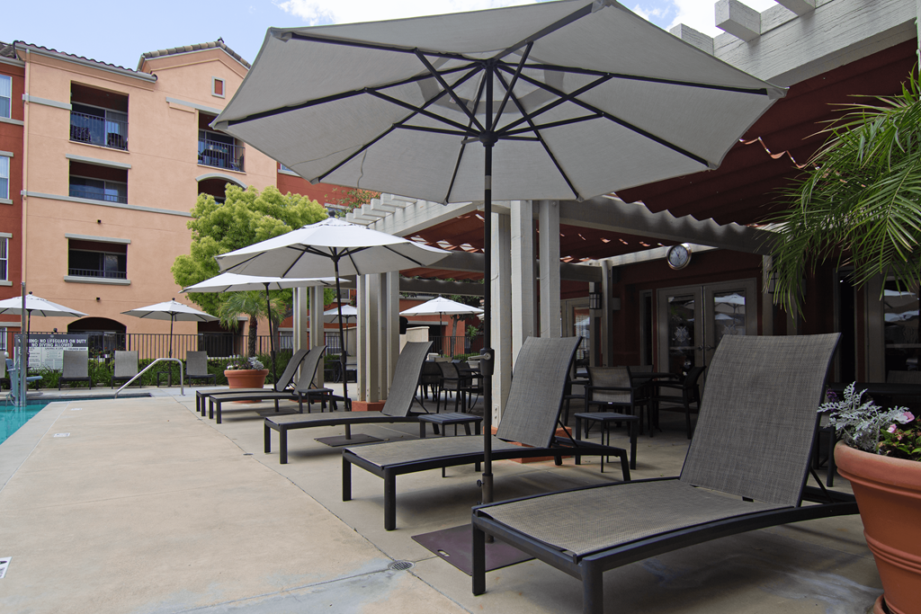 A patio with chairs and umbrellas is surrounded by a pool and buildings.