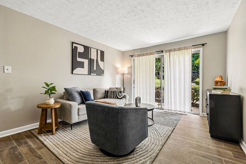 A living room with a grey sofa, a wooden side table, a rug, and a large window with curtains.