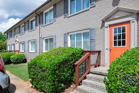 A grey building with a red door and a green bush in front.