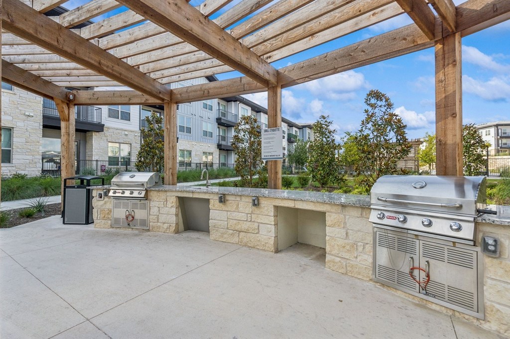 A BBQ grill is on a patio with a stone wall and a wooden pergola.