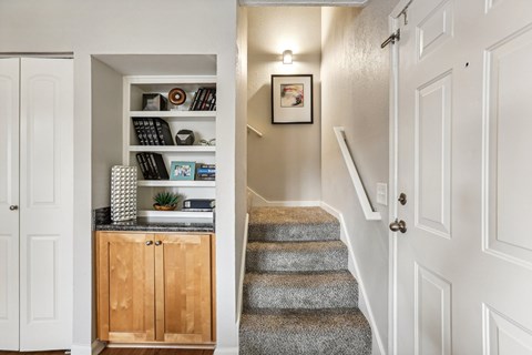 A hallway with a staircase and a cabinet with a shelf above it.