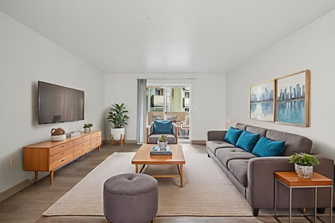 A living room with a grey ottoman, a brown coffee table, a grey couch, a brown side table, a wooden cabinet, a flat screen TV, a potted plant, and a painting on the wall.