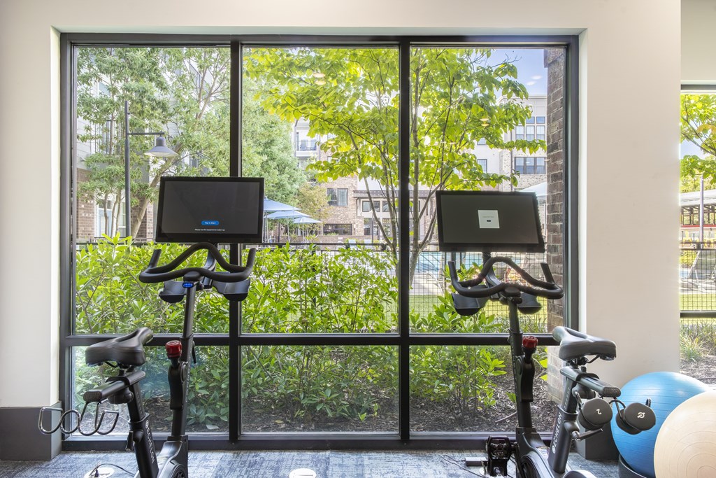Two exercise bikes are in front of a window with a view of trees and buildings outside.