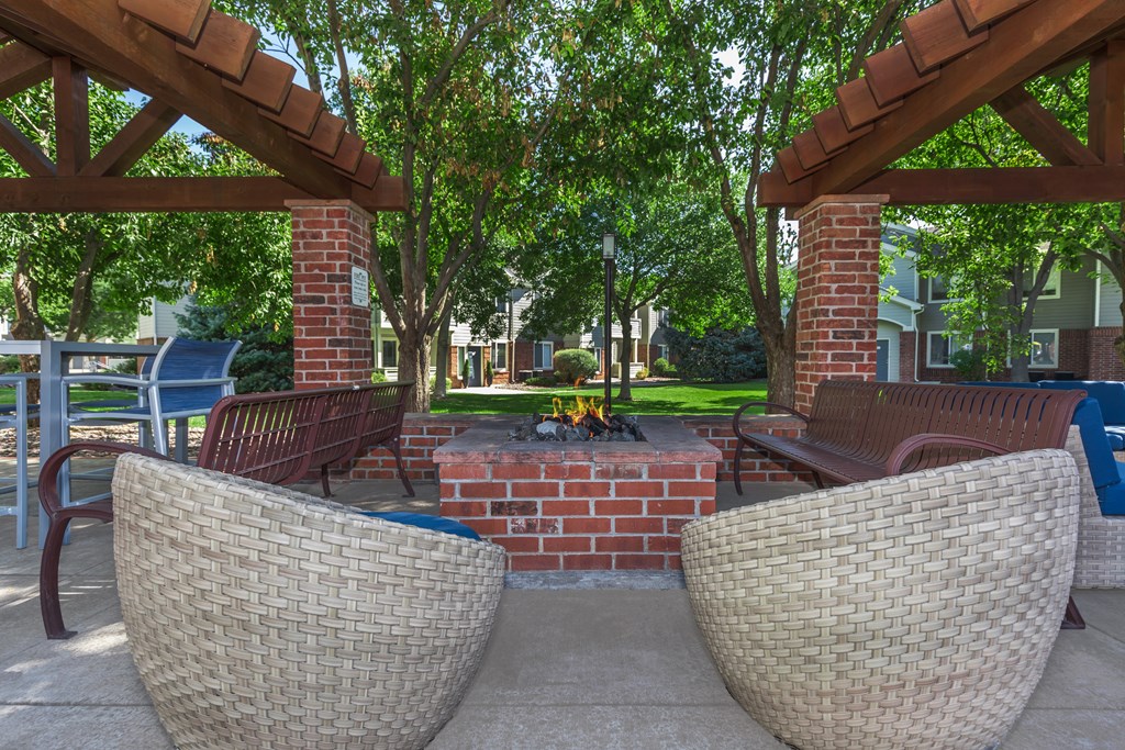 Two wicker chairs are placed under a wooden pergola with a firepit.