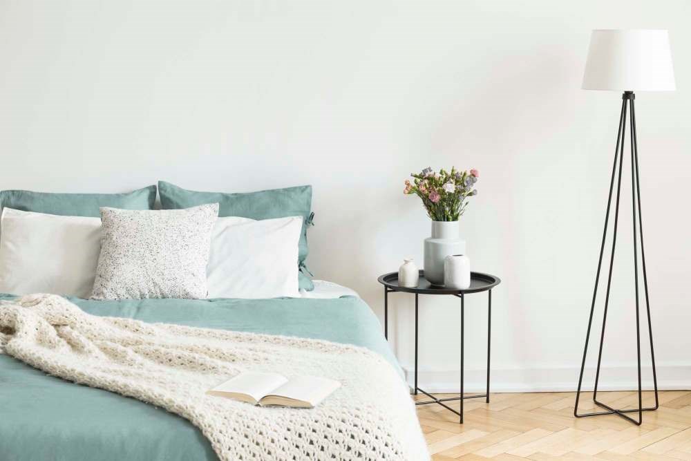 A cozy bedroom scene with a bed, a side table with a vase of flowers, a lamp, and a book.