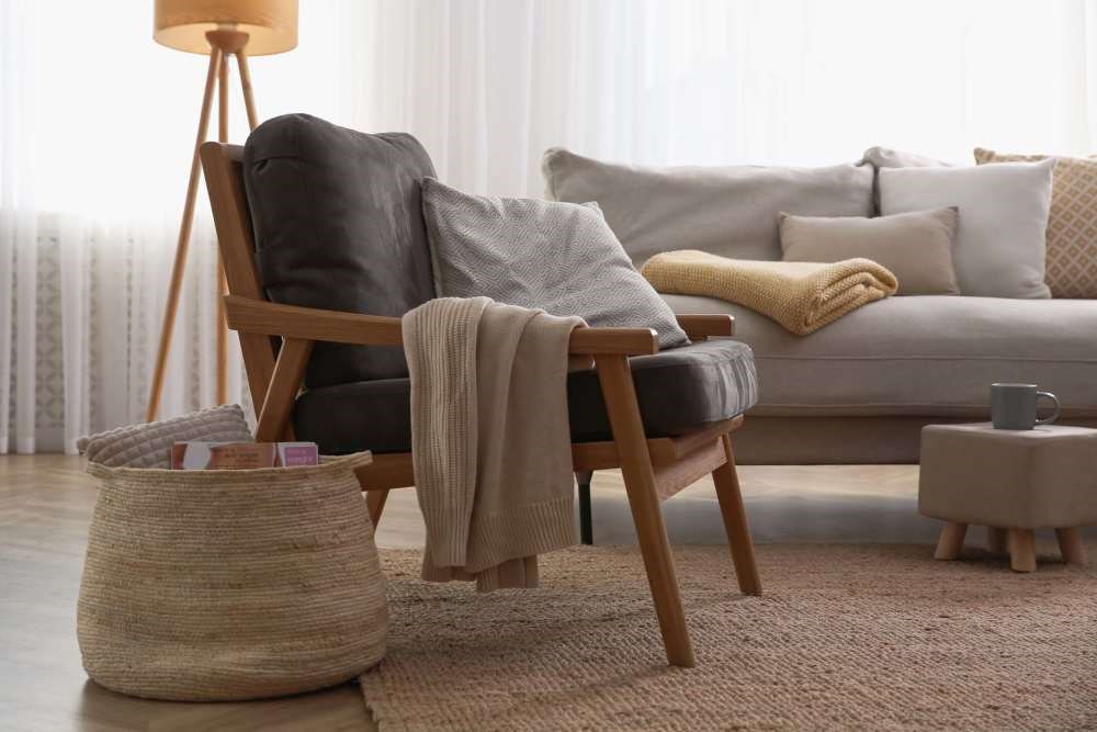 A cozy living room with a grey couch, a wooden chair with a grey cushion, a beige throw blanket, a small wooden side table, and a woven basket on the floor.