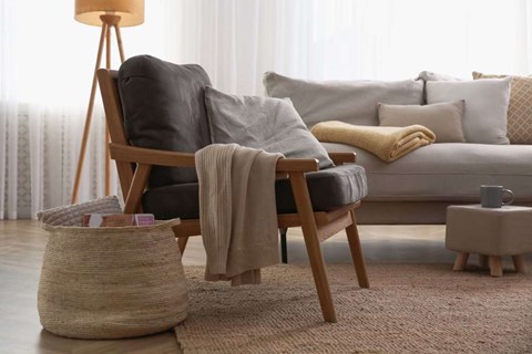 A cozy living room with a grey couch, a wooden chair with a grey cushion, a beige throw blanket, a small wooden side table, and a woven basket on the floor.