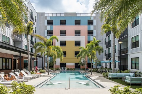 A swimming pool is surrounded by palm trees and lounge chairs in front of a colorful building.