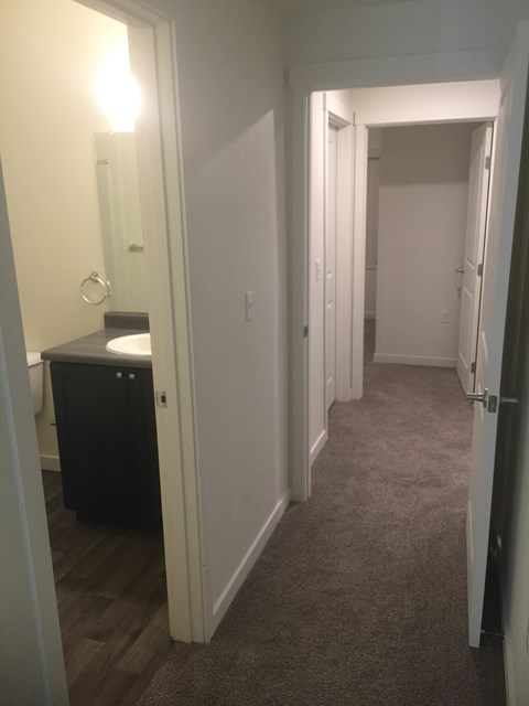 A hallway with a door on the left and a bathroom visible through the door.