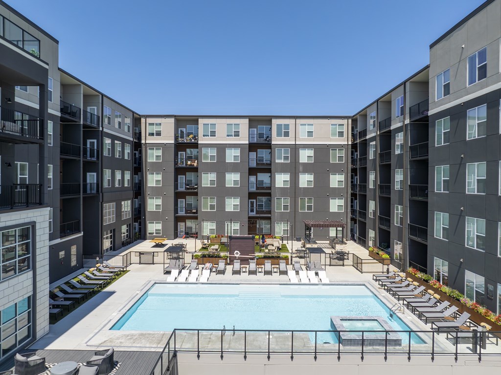 A large swimming pool surrounded by lounge chairs and umbrellas in the middle of apartment buildings.
