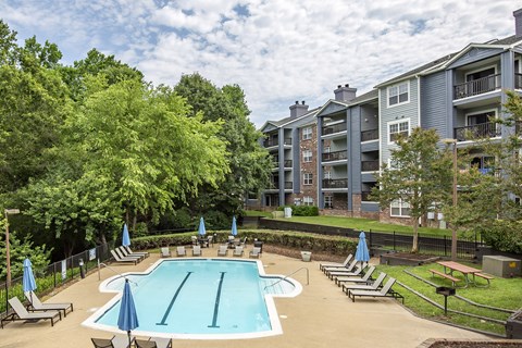 A swimming pool surrounded by trees and chairs with a building in the background.
