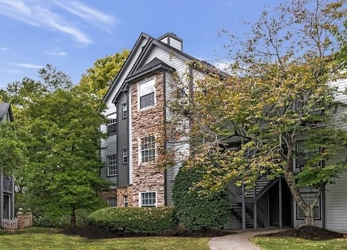 A house with a grey roof and a stone chimney is surrounded by green trees.