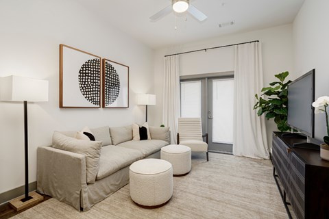 A living room with a grey couch, a white ottoman, a painting on the wall, a television, and a plant.
