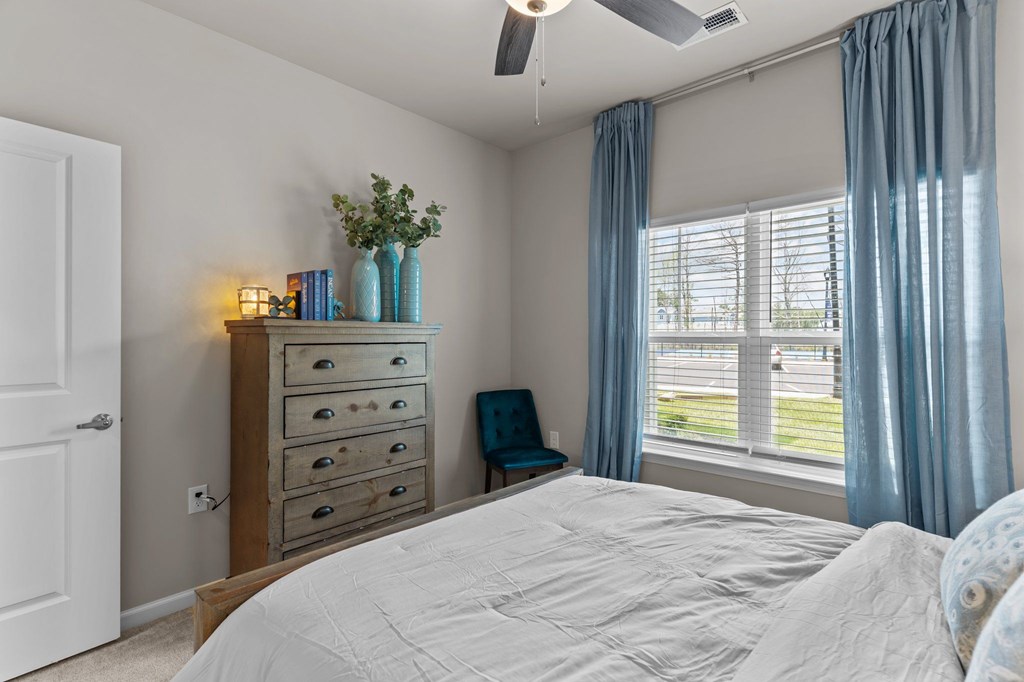 A bedroom with a large bed, a dresser with drawers, a window with blue curtains, and a ceiling fan.
