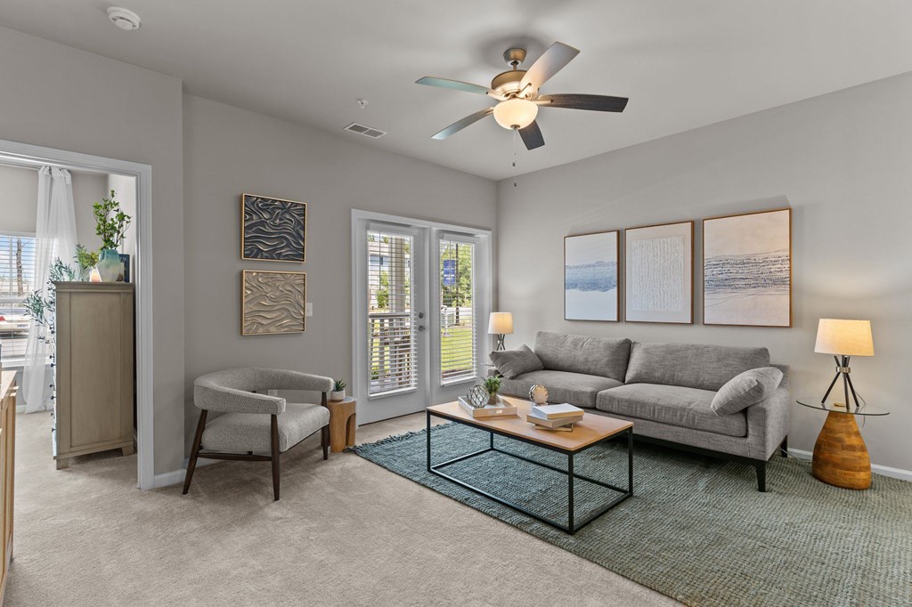 A living room with a grey couch, a chair, a table with a vase and a plant on it, and a ceiling fan.