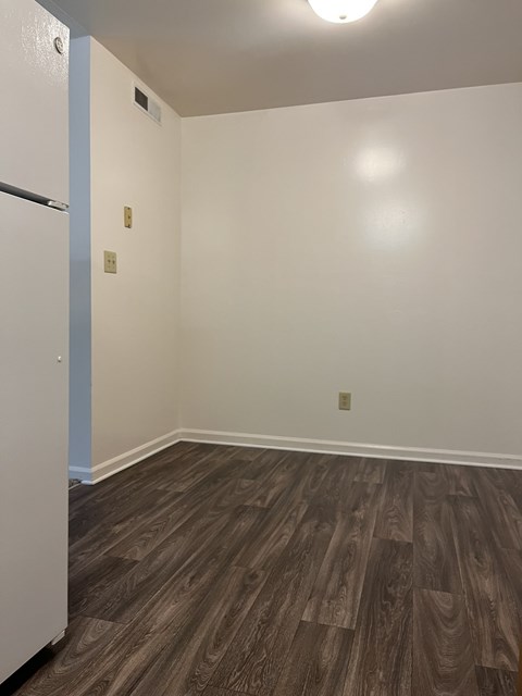 A room with a wooden floor and a white wall with a fridge on the left.