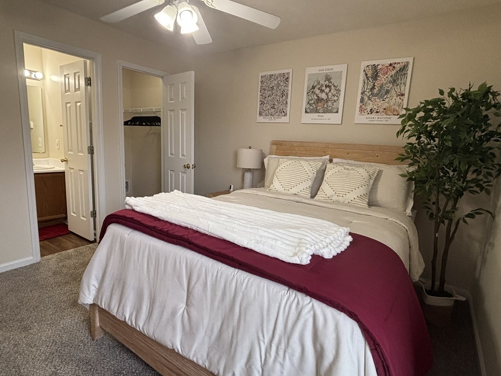 A bed with a white comforter and a red blanket is in the foreground of a room with a ceiling fan and a potted plant.