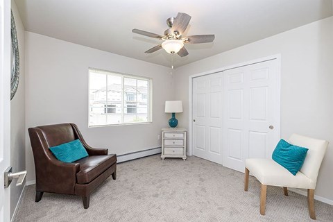 A room with a brown chair and a white chair, a ceiling fan, and a small table with a blue vase on it.