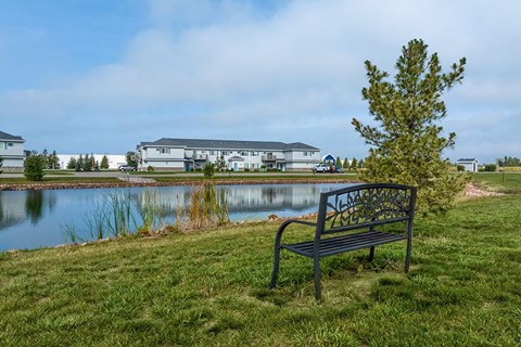 A bench with a metal armrest and a decorative design on the backrest is situated on a grassy area next to a body of water.