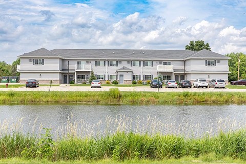 A large white building with a grey roof is situated by a body of water with greenery in the foreground.