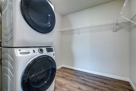 A white front load washing machine in a laundry room with a white wall and a shelf.