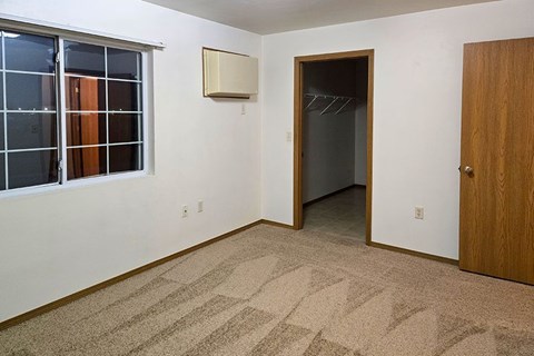 A room with a carpeted floor, a door, a window with blinds, and a wall-mounted air conditioning unit.