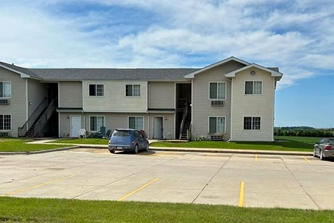 A grey car is parked in a parking lot in front of a two-story apartment building.