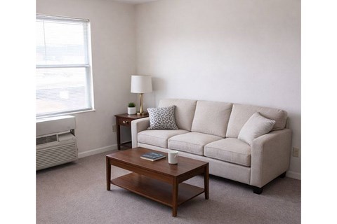 A living room with a beige couch, a small table with a lamp, and a window with an air conditioner.
