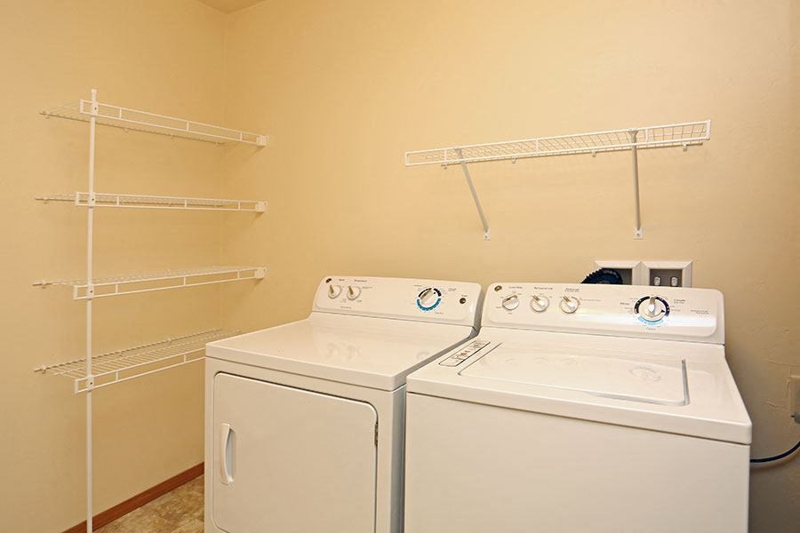 Two white washing machines in a laundry room with shelving above.