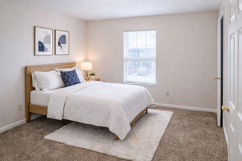 A bedroom with a bed, two pictures on the wall, a nightstand with a lamp, and a window.