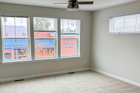 A room with a carpet floor, a ceiling fan, and a window showing a view of a red building.