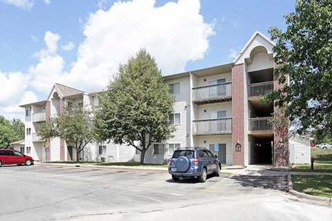A red car is parked in front of a white building with a balcony.