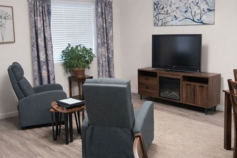 A living room with a grey couch, a grey chair, a grey armchair, a wooden table, a television, a plant, and a painting on the wall.