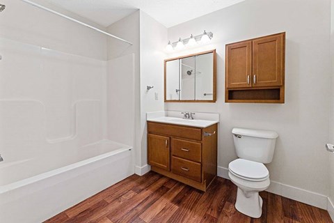 A white toilet sits next to a wooden cabinet and a white bathtub in a bathroom.