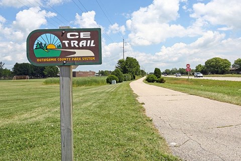 A sign for the CE Trail stands on a grassy area beside a paved road.
