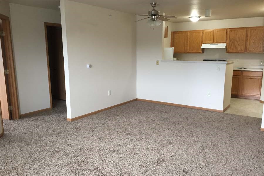 A room with a ceiling fan and a kitchenette in the background.