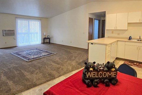 A living room with a rug, a table with a sign that says "welcome" and a kitchen area.