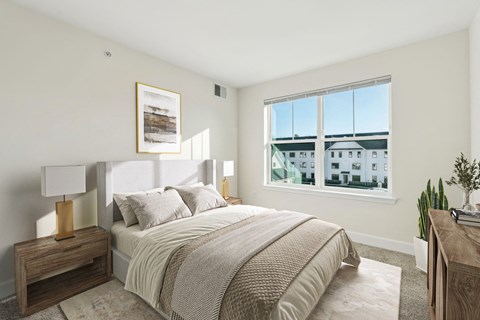 A bedroom with a large bed, a nightstand with a lamp, and a window overlooking a building.