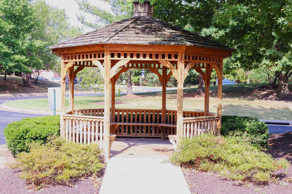 A wooden gazebo with a brown roof is surrounded by green bushes and trees.