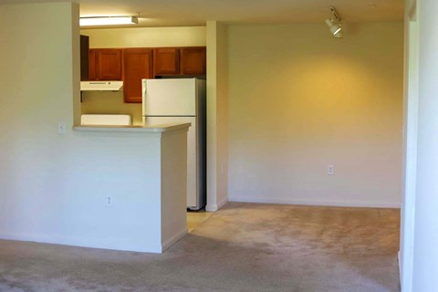 an empty living room with a kitchen with a refrigerator