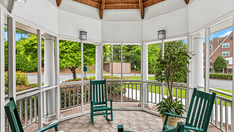 A white gazebo with a wooden roof and a white railing with green chairs and a tree in a pot.